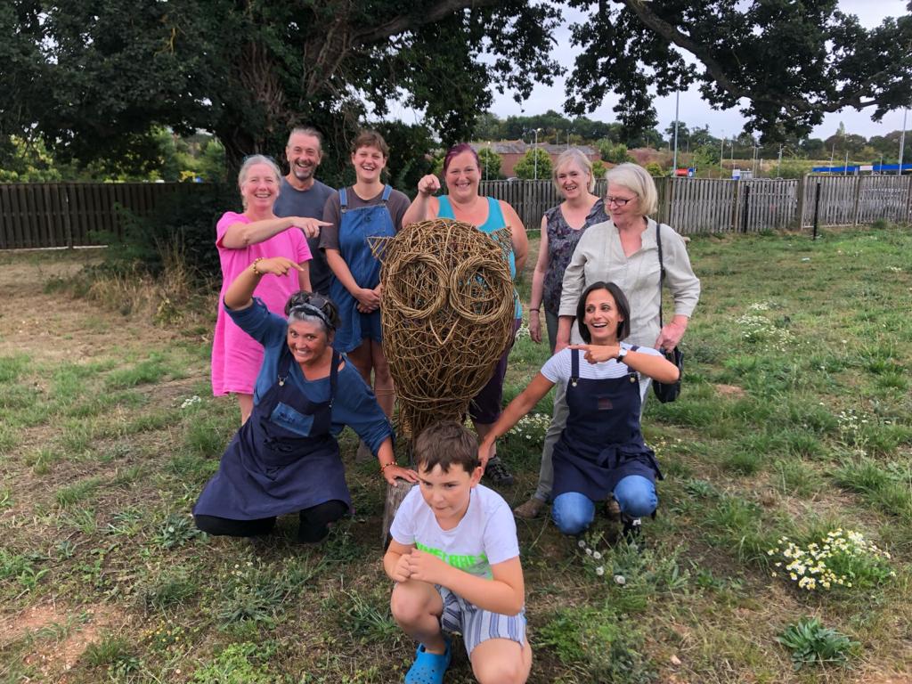 Weird Sticks workshop participants posing with finished willow owl sculpture in Community Garden at Crediton Heart Project