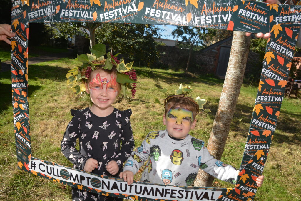 Two children wearing their willow harvest crowns made with Weird Sticks pose with a selfie frame at the Cullompton Autumn Festival