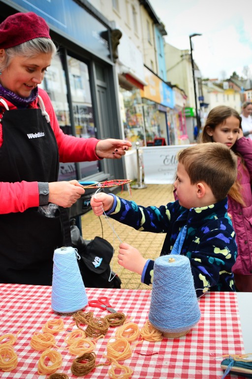 Vik from Weird Sticks helping smiling child to make willow and wool fish in Brixham at Playing in the Streets Easter 2023