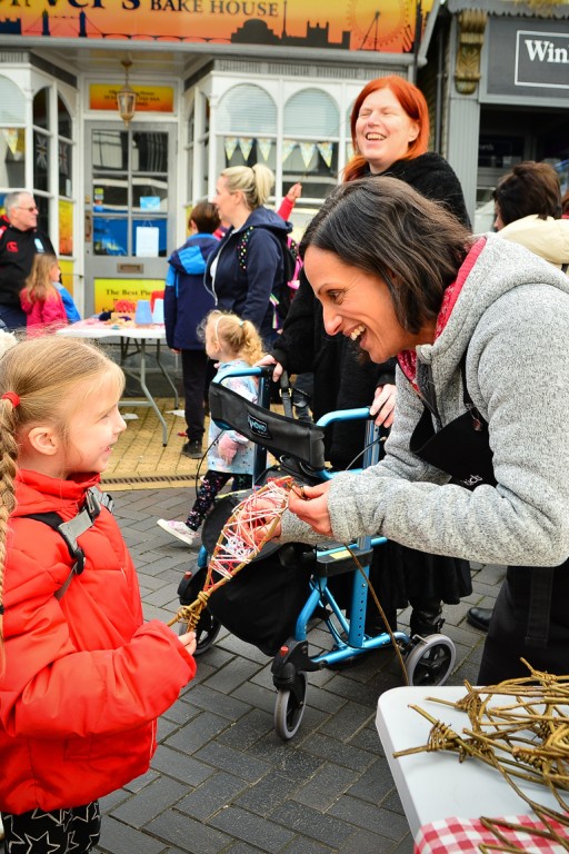 Hannah from Weird Sticks helping smiling child to make willow and wool fish in Brixham at Playing in the Streets Easter 2023