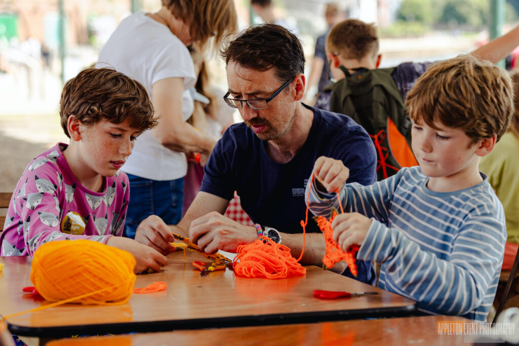 Parent and two children weaving a spinning sun at the Weird Sticks workshop, Exeter Quay